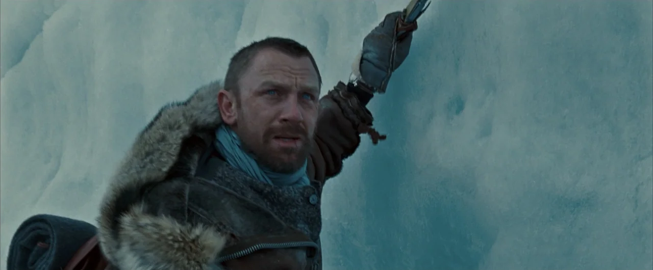 The Golden Compass (2007) movie still: medium — A man in a thick fur parka scales a sheer wall of blue ice. He reaches upward with one hand while…