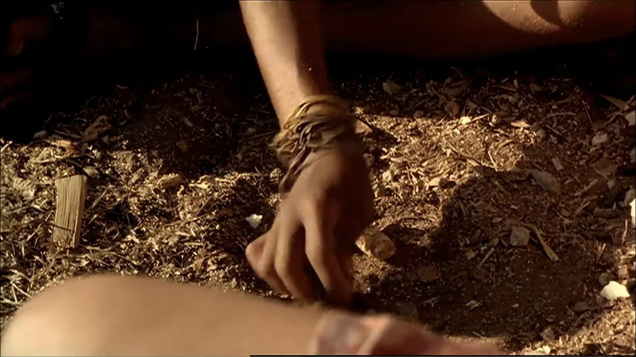 In the Middle of the World (1990) movie still: closeup — A patch of dry, sun-drenched earth is littered with wood shavings and small stones. A human…