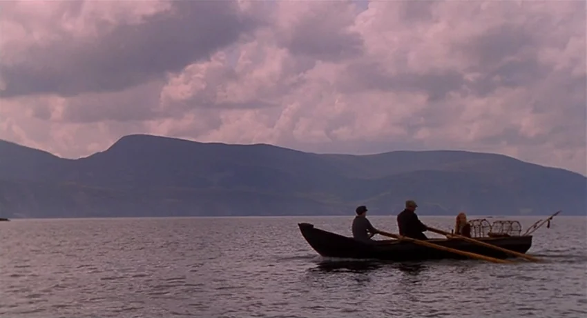 The Secret Of Roan Inish (1994) movie still: wide — A calm expanse of water lies beneath a large, cloudy mountain range. Three individuals in a…