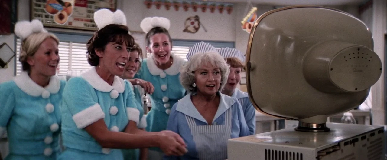 Grease (1978) movie still: medium — A group of six waitresses in matching teal uniforms watch a small television monitor. They stand behind a…
