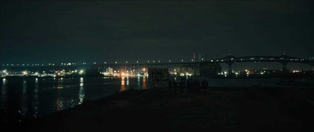 Guns Up (2025) movie still: wide — A dark riverbank at night overlooks a large steel bridge spanning the water. A cluster of people stands near the…