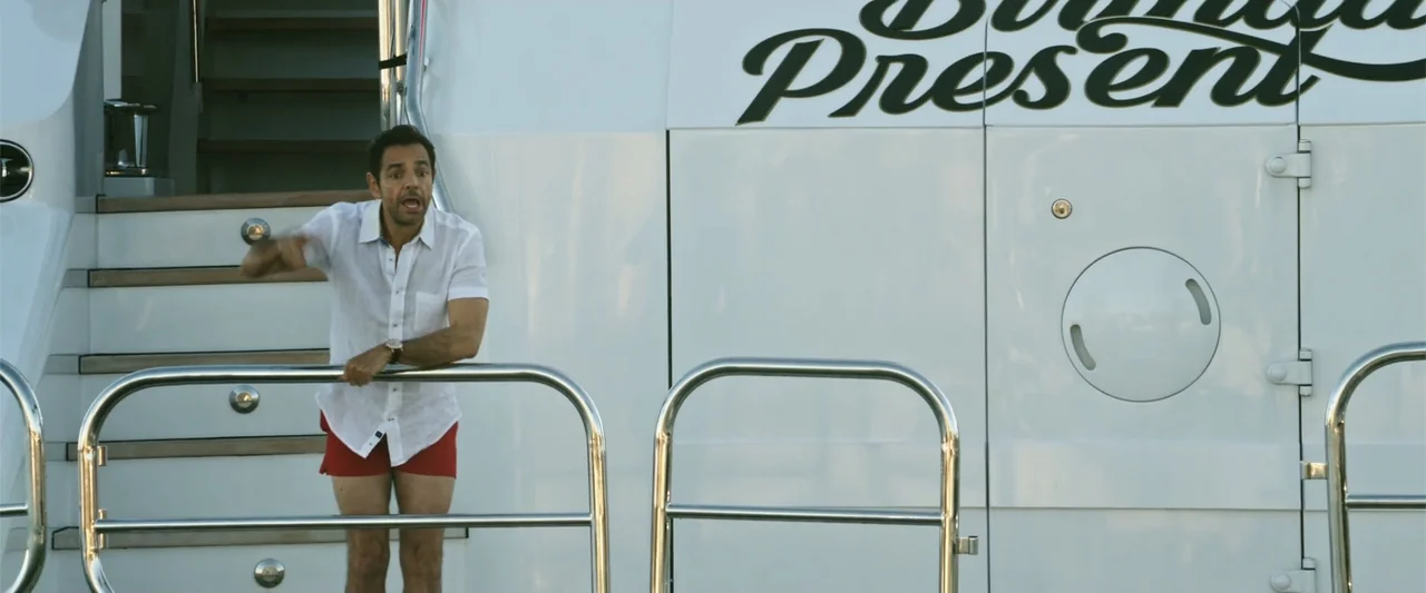 Overboard (2018) movie still: medium — A man stands on the white exterior deck of a large yacht. He leans against a metal railing while gesturing…
