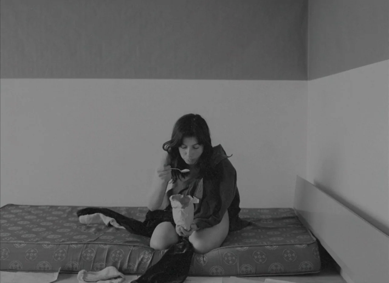 Je Tu Il Elle (1974) movie still: medium — A plain bedroom features a thin mattress on the floor against a two-tone wall. A young woman in a dark…