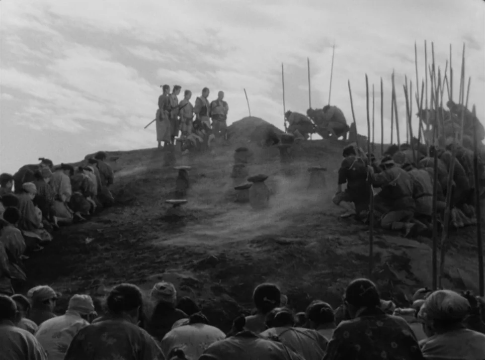 Seven Samurai (1954) movie still: wide — A large crowd of people gathers on a grassy slope under an overcast sky. The individuals are kneeling in…