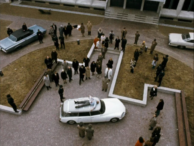 Making of: World On A Wire (1973) movie still: establishing — A stone courtyard features angular planters and a large crowd surrounding a white…