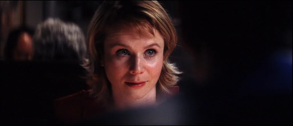 Punch Drunk Love (2002) movie still: closeup — A dimly lit indoor space with blurred figures in the dark background. A woman sits in a booth looking…