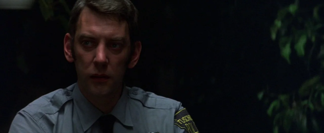 Klute (1971) movie still: medium — A dimly lit room serves as the setting for a professional interaction. A man in a police uniform stares forward…
