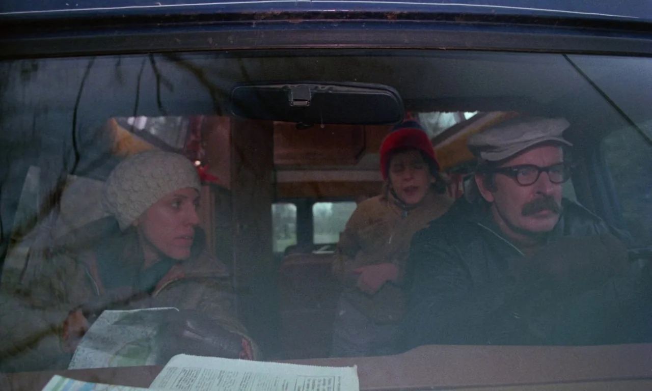 Rabid (1977) movie still: medium — The interior of a moving van features a family of three during a daytime trip. The father drives while the mother…