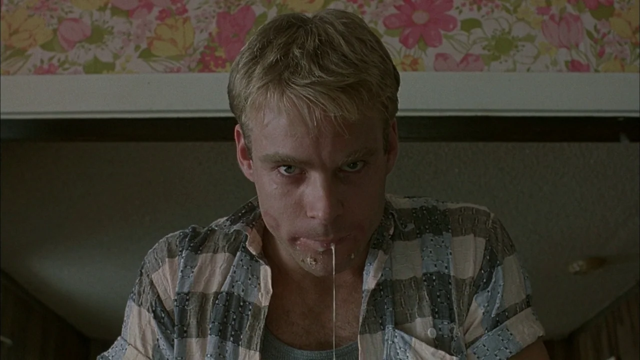 Whore (1991) movie still: closeup — An interior space features floral patterned wallpaper behind a man's head. The man stares directly at the camera…
