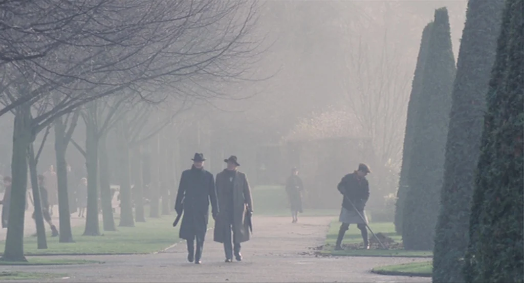 The King’s Speech (2010) movie still: wide — A foggy public park features rows of mature trees and tall shaped topiary hedges. Two men in formal…