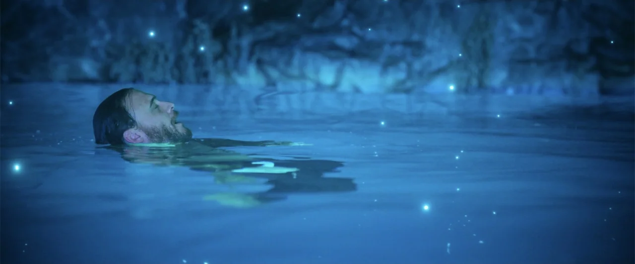 Blue Cave (2024) movie still: medium — A man drifts in a dark pool surrounded by a textured rock wall. Glowing light particles float in the air…