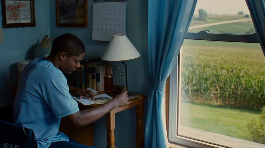 Sugar (2008) movie still: medium — A quiet room has light blue walls decorated with framed prints and a calendar. A young man sits at a wooden desk…