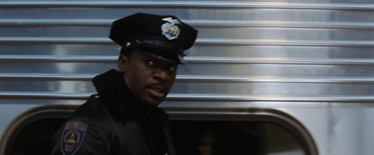 Fallen (2004) movie still: medium — A man in a police uniform stands before the corrugated aluminum side of a train. The officer looks toward the…