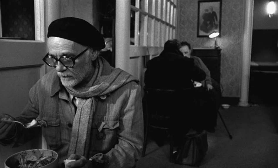 The Man From London (2007) movie still: medium — An interior space with patterned wallpaper, framed artwork, and wooden partitions. A man in a beret…