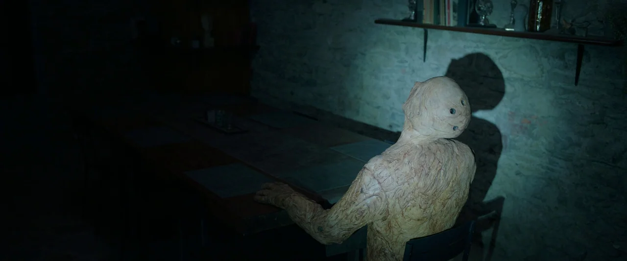 Oddity (2024) movie still: medium — A dark dining room with a rough stone wall background and a wooden table. A creature with textured skin sits in…