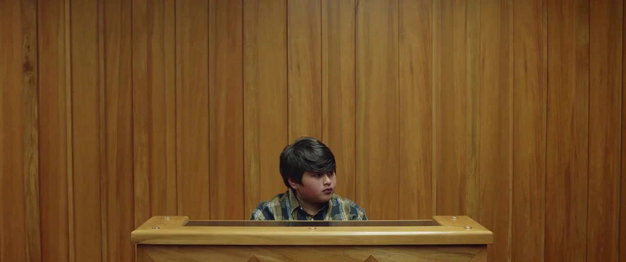 Hunt for the Wilderpeople (2016) movie still: medium — A young boy sits behind a polished light-wood altar against a matching paneled wall. The…