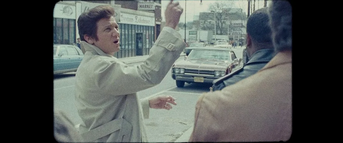 American Hustle (2013) movie still: medium — A man in a trench coat stands on a city street near parked cars. He gestures with his hand towards the…