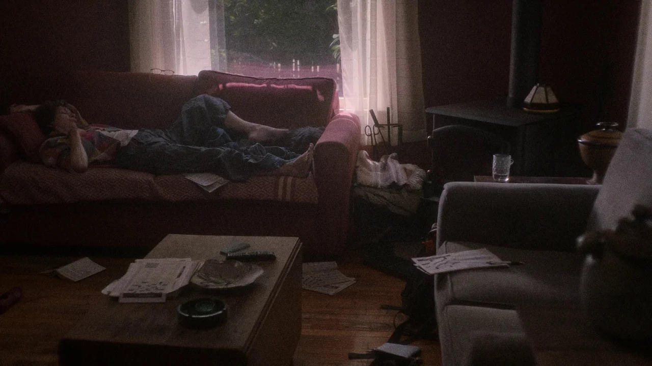 Showing Up (2022) movie still: wide — A dimly lit room with a red fabric sofa, wooden floors, and a coffee table. A person lies on the sofa, resting…