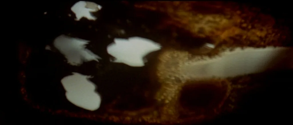 Two Lane Blacktop (1971) movie still: closeup — A dark, viscous liquid fills the frame with swirling patterns of a lighter fluid. The light reflects…