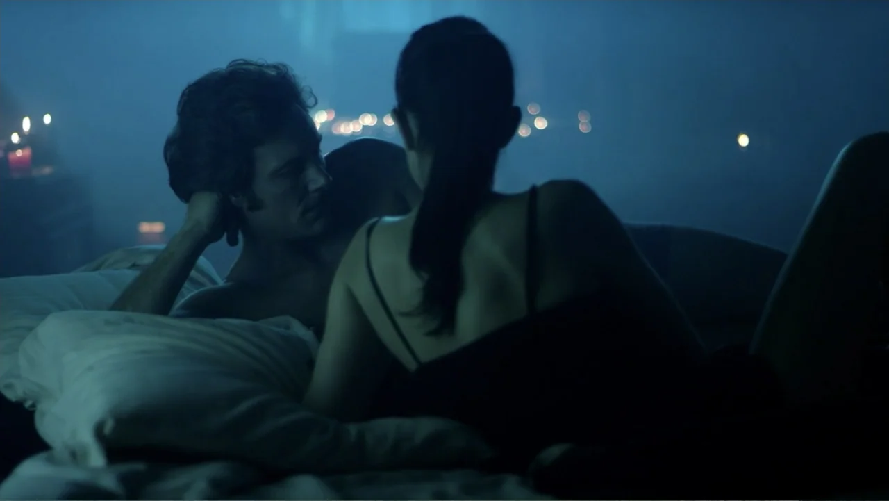 Entre las sombras (2025) movie still: medium — A dark bedroom interior is illuminated by soft blue light and distant bokeh. A man and a woman lie in…