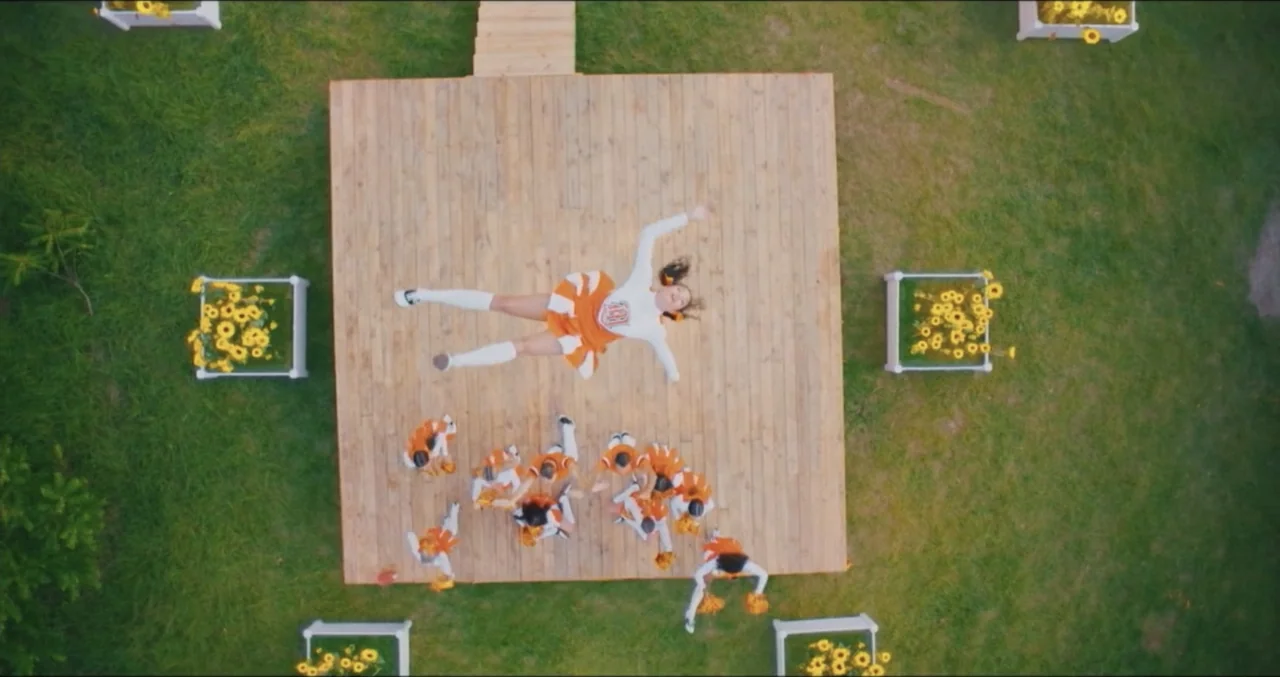 Eye for an Eye (2025) movie still: wide — A wooden platform sits on a green lawn surrounded by square planters of yellow flowers. A cheerleader in…