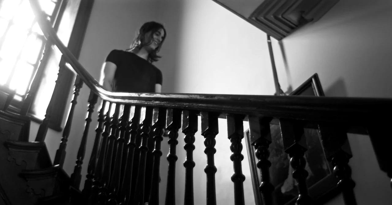 T Blockers (2023) movie still: medium — A wooden staircase with dark balusters leads toward a bright window at the top. A young man with long dark…