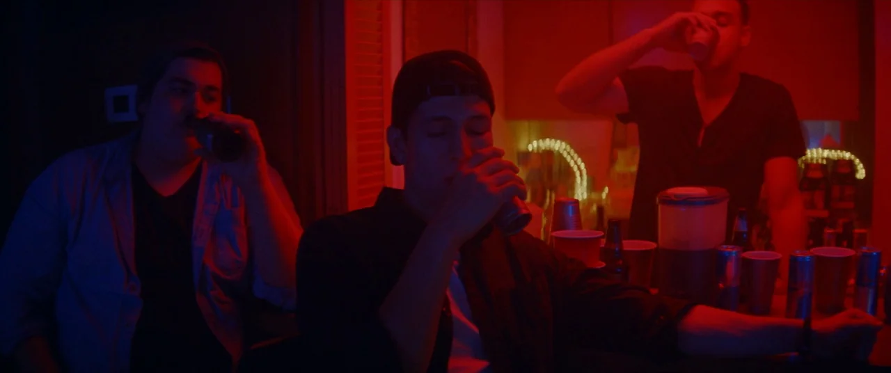 Assassination Nation (2018) movie still: medium — A dimly lit room bathed in harsh red neon light during a social gathering. Three young men stand…