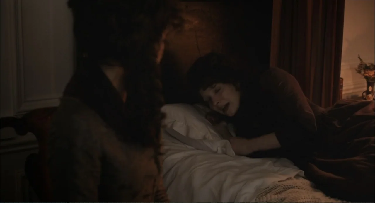 Love & Friendship (2016) movie still: medium — A dark bedroom with dark wooden furniture and a person lying in a bed. One woman sits in the…