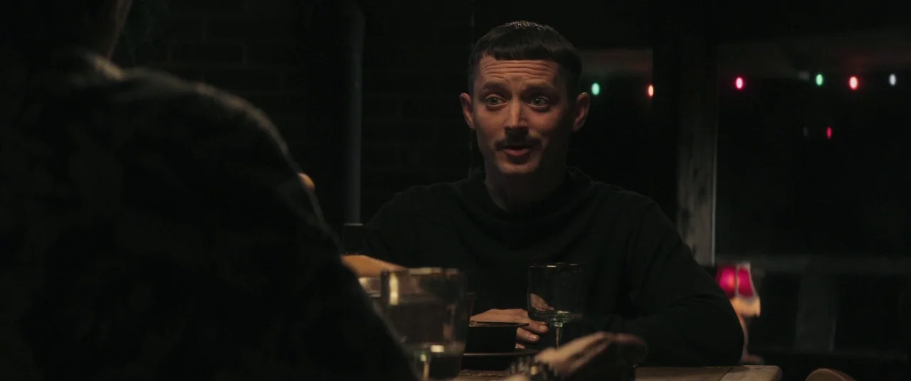 Come To Daddy (2019) movie still: medium — A dark indoor space with a wooden table and blurred festive lights in background. A man with a dark…