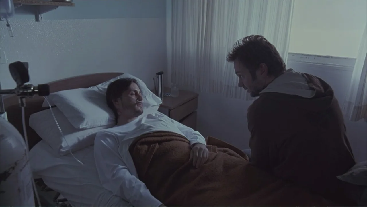 A Distant Place (2020) movie still: medium — A spartan hospital room features a medical bed against a window with thin curtains. A man in a hoodie…
