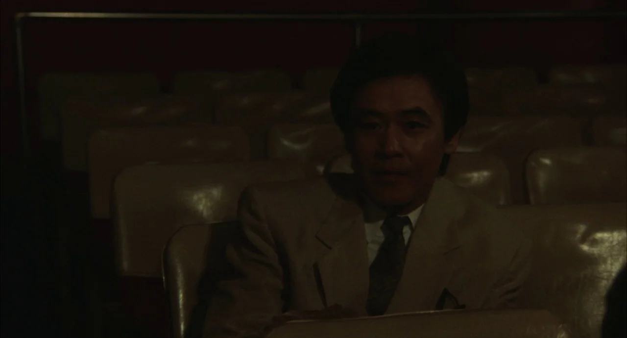 The Discarnates (1988) movie still: medium — A man sits in the dim interior of a movie theater with empty seats. The man looks ahead while sitting…