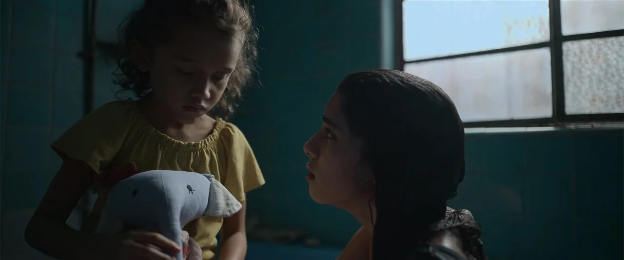 Evil Eye (2020) movie still: medium — A dimly lit tiled interior space features a window with daylight filtering through. A young woman and a small…