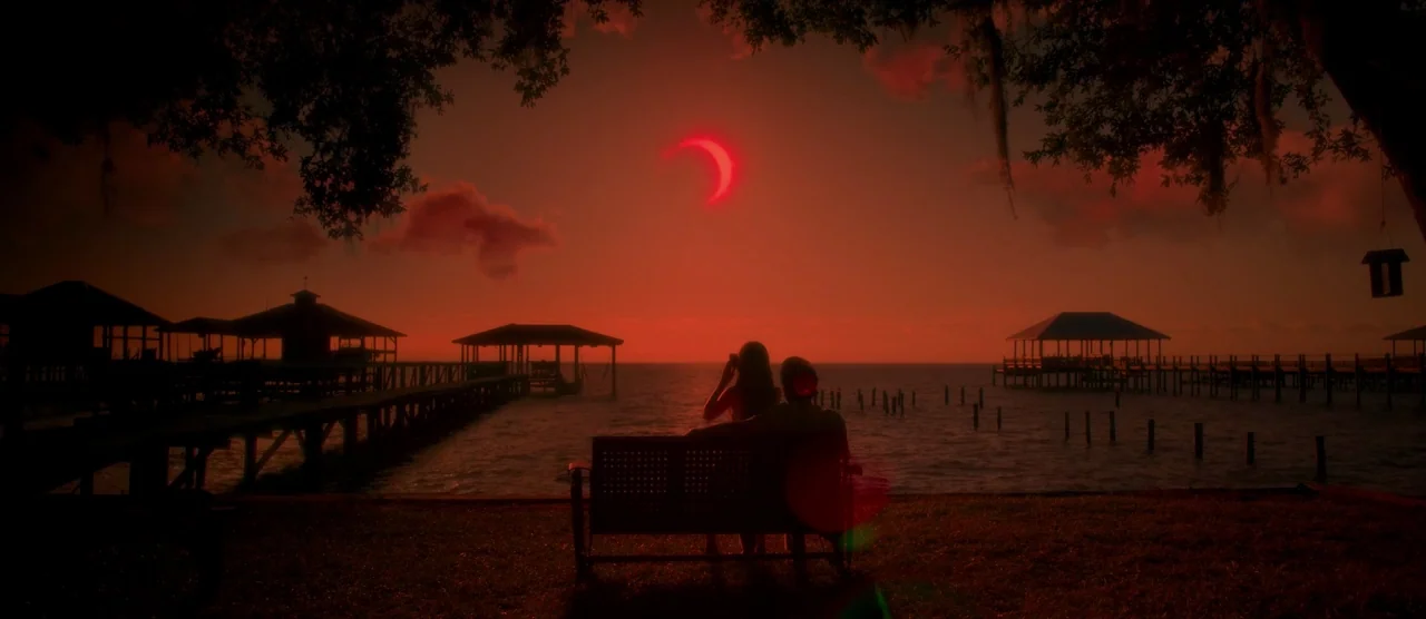 Gerald’s Game (2017) movie still: wide — A dark wooden pier extends into the water under a sky with a red eclipse. Two people sit together on a…