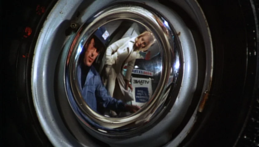 A Bay of Blood (1971) movie still: closeup — A night scene shows a reflection in a car hubcap at a gas station. A man in a work jacket and a woman…