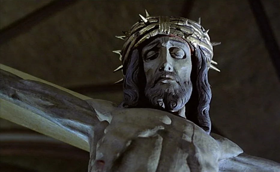 Possession (1981) movie still: closeup — A carved wooden statue of a crucified figure hangs in a dark space. The figure wears a crown of thorns and…