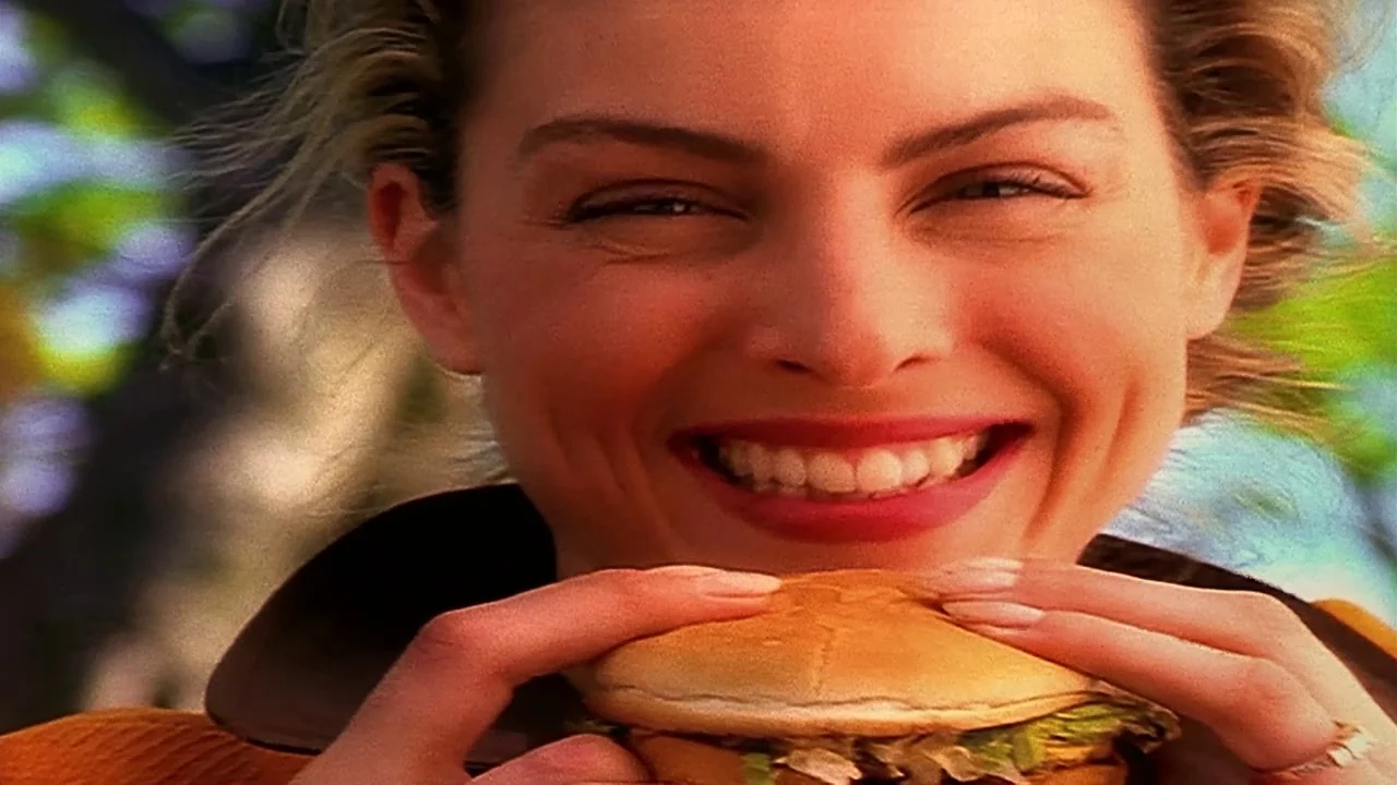 Naqoyquatsi (2002) movie still: closeup — A woman stands in a bright, sunlit outdoor area with blurred foliage behind her. She holds a cheeseburger…
