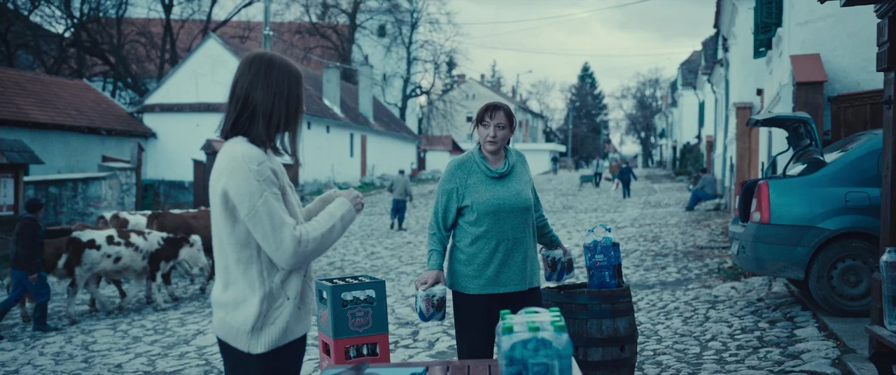 R.M.N. (2022) movie still: medium — A European cobblestone village street with white-walled houses and a hatchback car parked nearby. Two women…