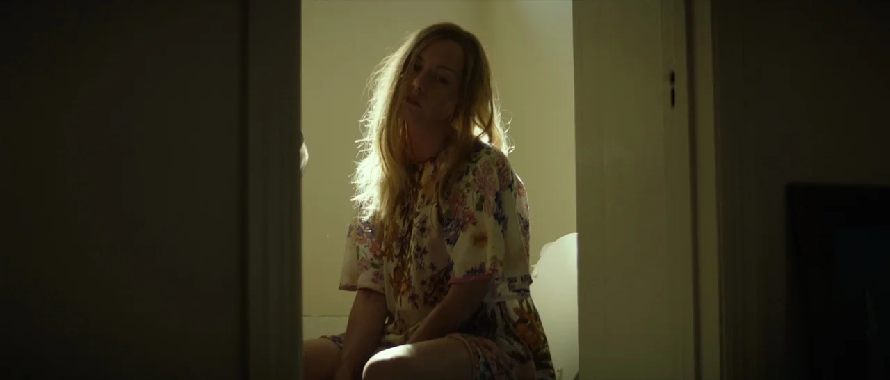 Ingrid Goes West (2017) movie still: medium — A dimly lit room with walls painted in a light beige tone. A woman with disheveled hair sits on a…