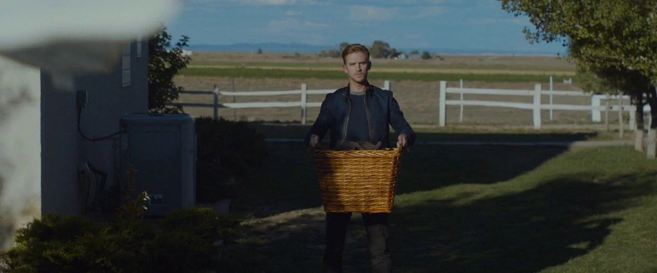 The Guest (2014) movie still: fullbody — A man stands on a grassy lawn in front of a rural house. He holds a large wicker basket with both hands…