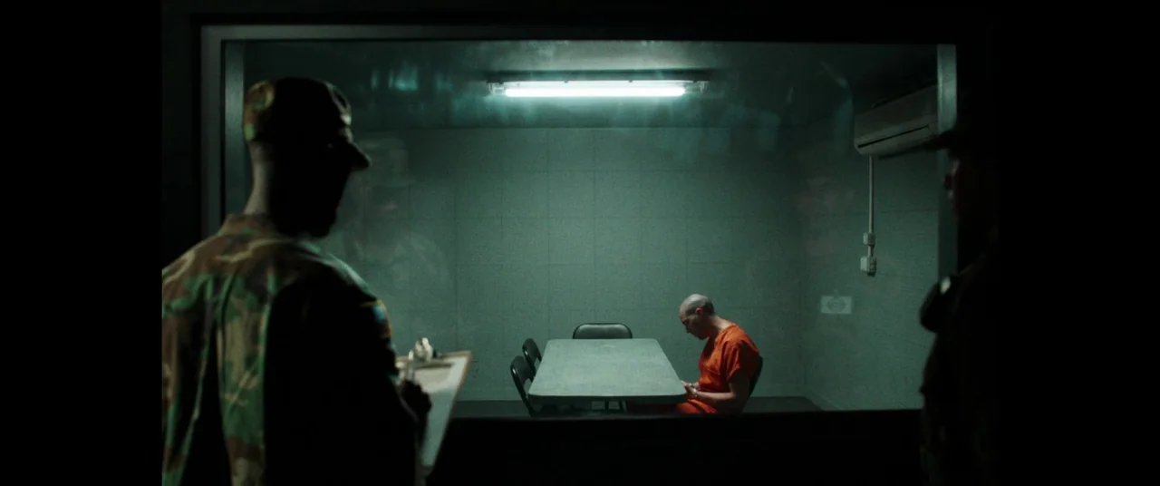 The Mauritanian (2021) movie still: wide — An observation room looks through a glass window into a sterile green-walled room. A lone man in an…