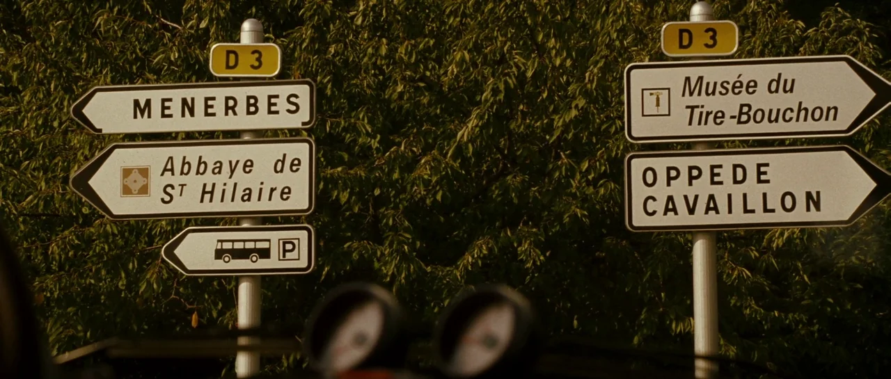 A Good Year (2006) movie still: medium — Two French road signposts stand against a dense backdrop of green deciduous trees. The signs direct traffic…