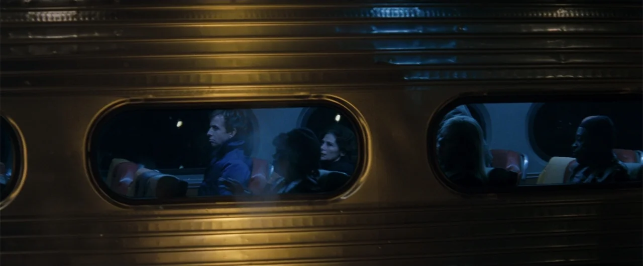 Fallen (2004) movie still: wide — The exterior side of a stainless steel passenger train features two illuminated oval windows at night. Multiple…