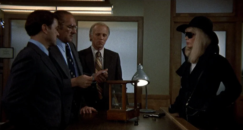 Family Plot (1976) movie still: medium — A dimly lit room with wood-paneled walls and glass partitions separating workspace areas. Three men in…