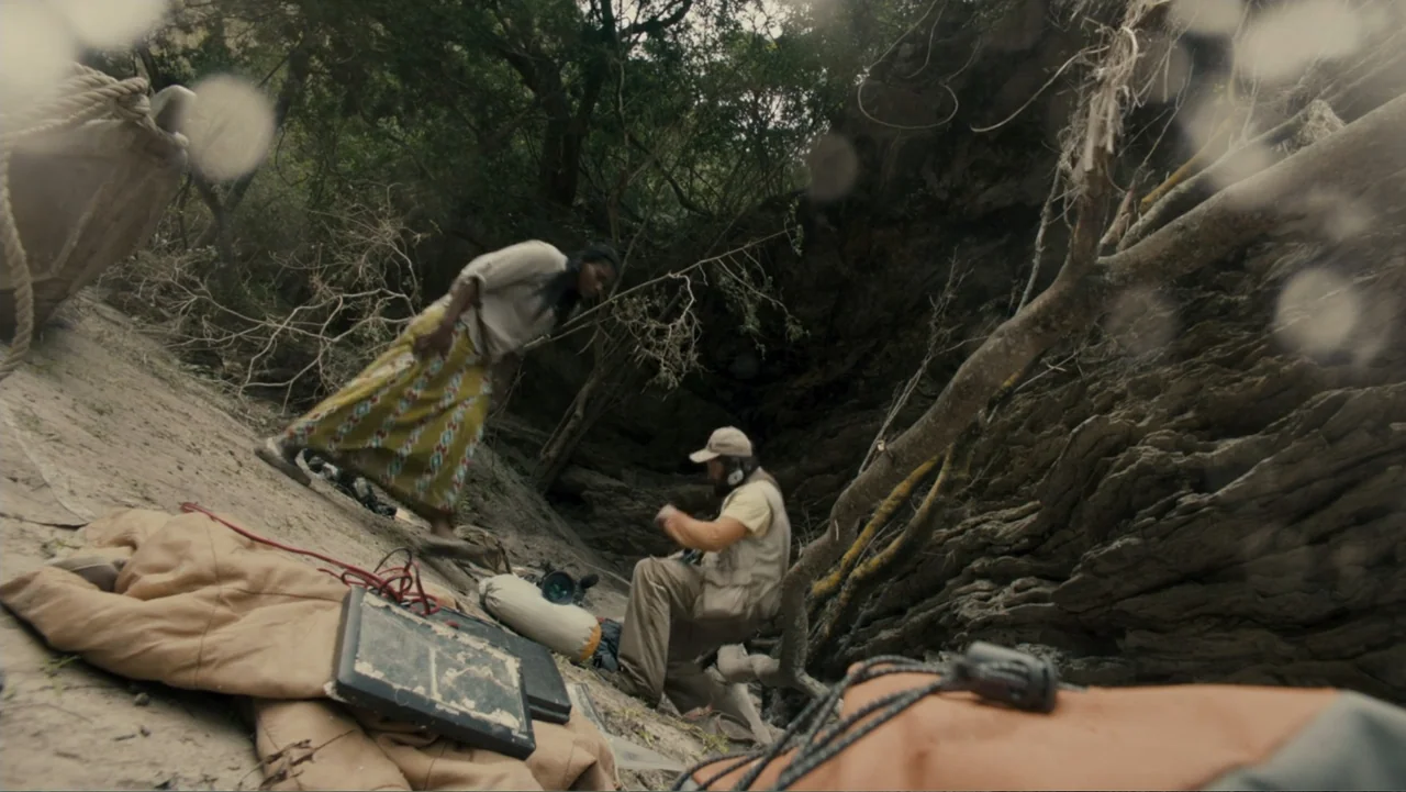 The Dinosaur Project (2012) movie still: fullbody — A rocky ravine floor features dense foliage and scattered camera equipment on the ground. A…