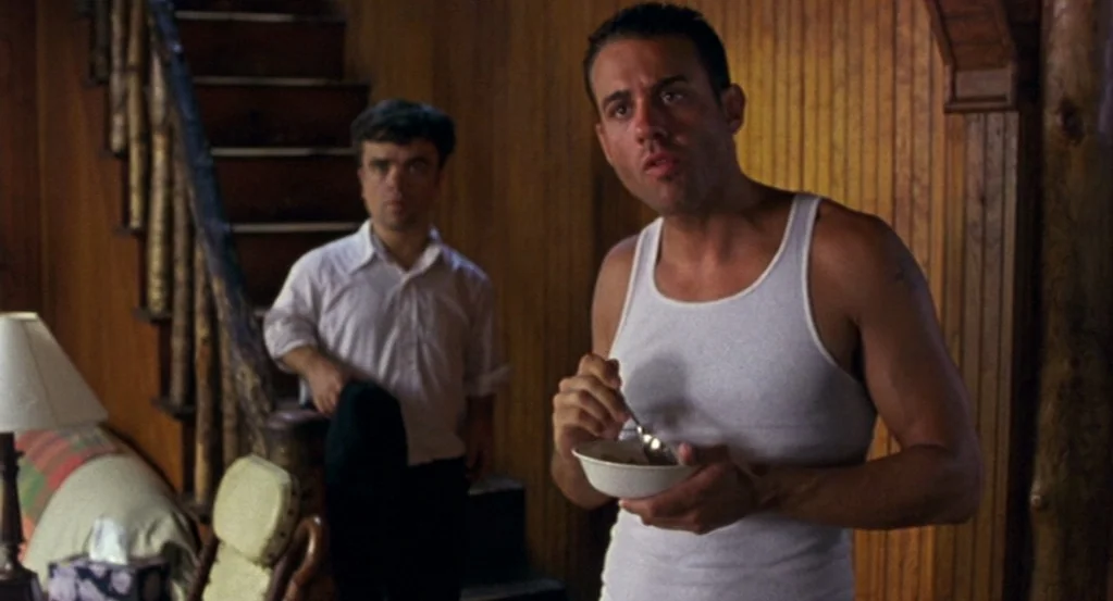 The Station Agent (2003) movie still: medium — A room features vertical wood paneling and a dark staircase in the background. One man holds a bowl…