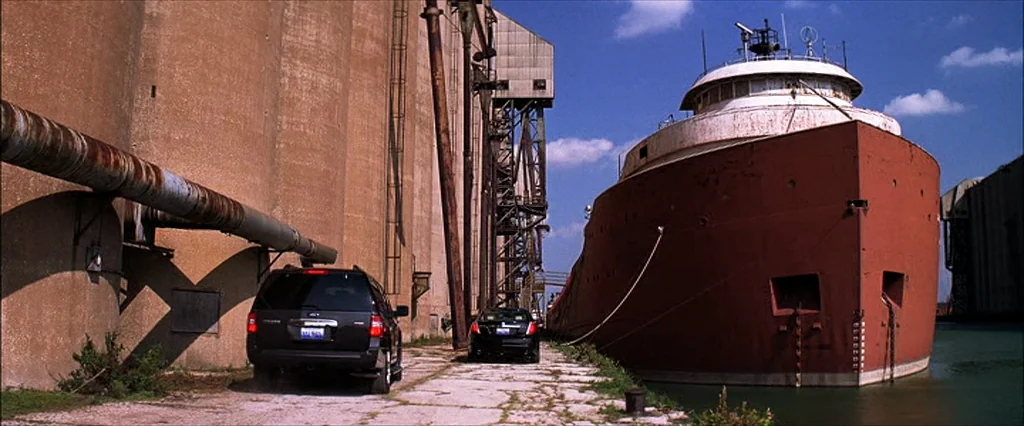 The Dark Knight (2008) movie still: wide — A rusted cargo ship sits docked beside a massive, weathered concrete grain elevator structure. Two modern…