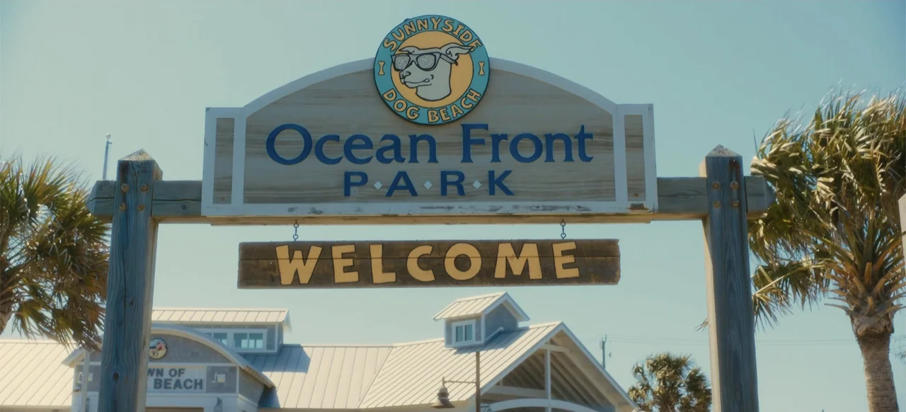 Merv (2025) movie still: wide — A wooden park entrance sign displays the name Ocean Front Park under bright sun. Palm trees flank the support beams…