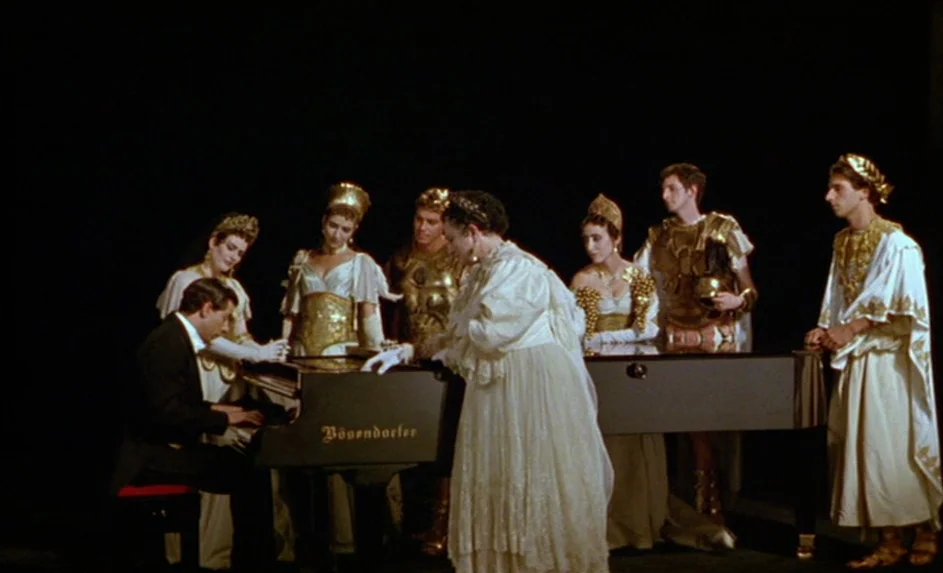 Wittgenstein (1993) movie still: wide — A group of people dressed in historical Roman attire stand on a dark stage. A man in a suit plays a black…