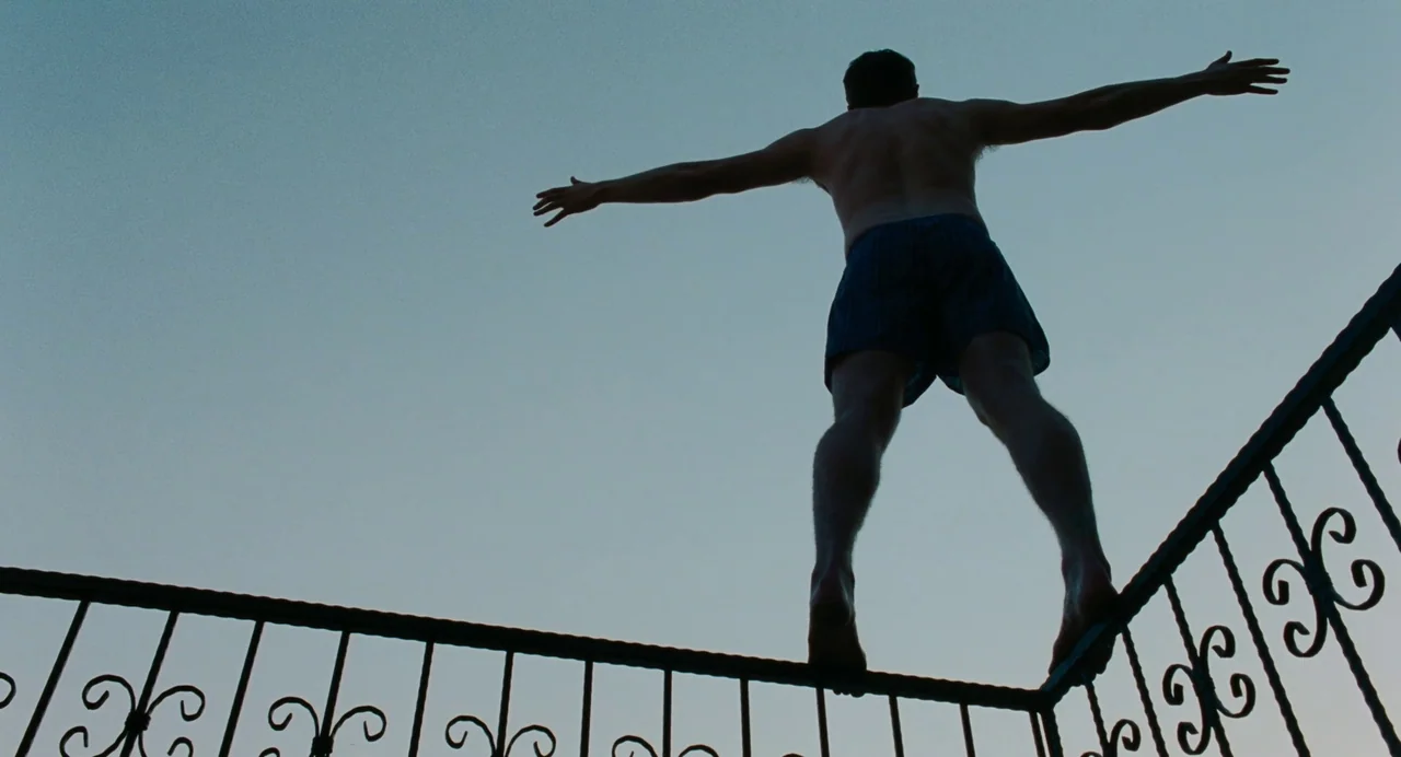 Aftersun (2022) movie still: fullbody — A man stands atop a decorative metal railing against an open blue sky. He maintains his balance with arms…