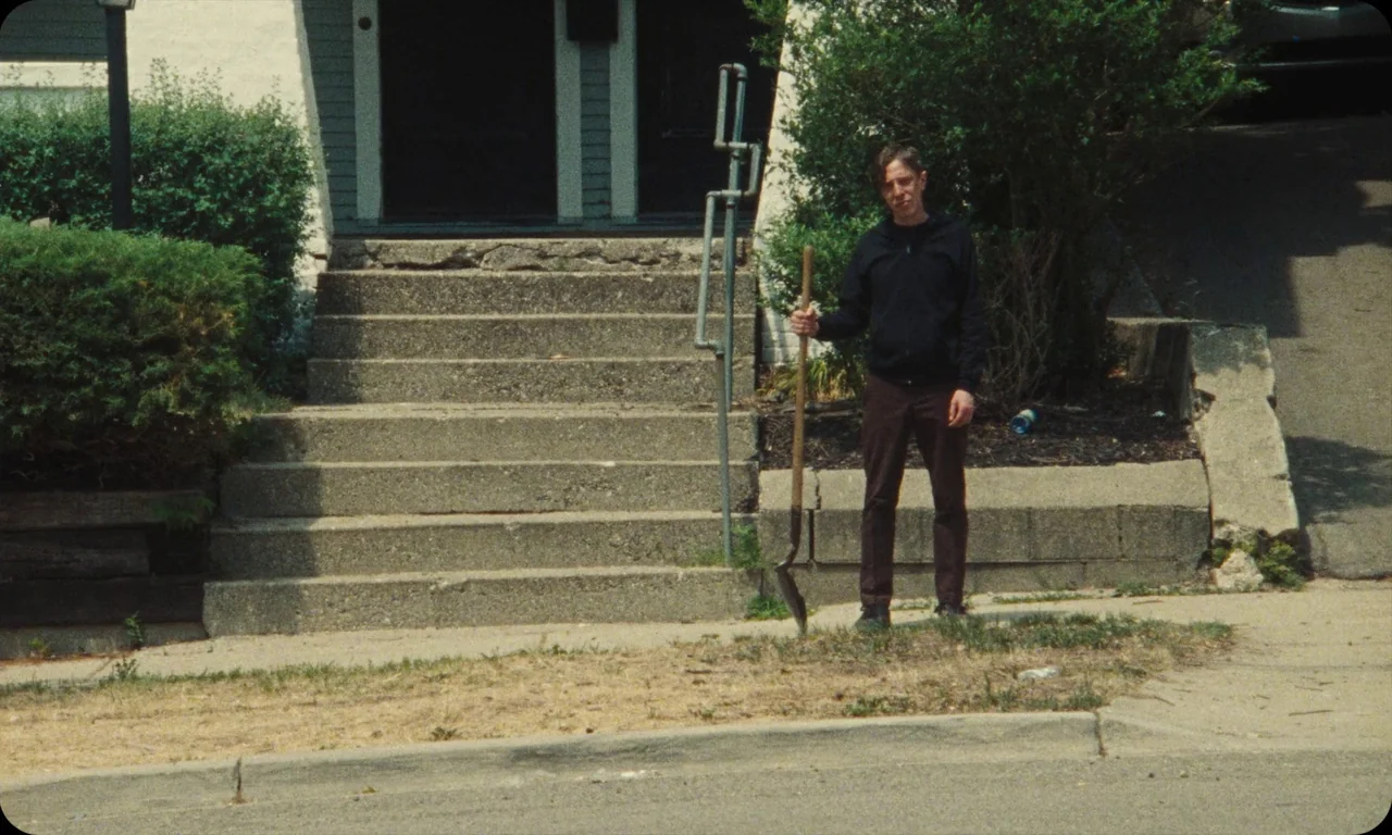Vulcanizadora (2024) movie still: fullbody — A man stands on a residential sidewalk in front of concrete building entrance steps. He holds a…